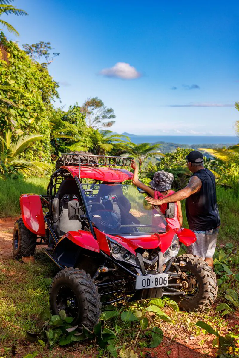 Terra Trek Quad Bike Safari Two people stand by a red adventure buggy on a dirt trail, with lush tropical foliage and ocean views in Pacific Harbour, Fiji.