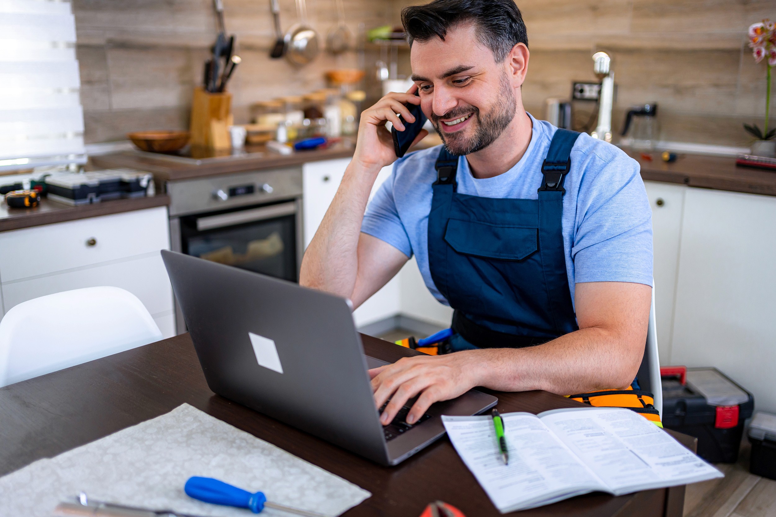 Handyman sits at a table in a kitchen, making phone calls and managing repair tasks online.