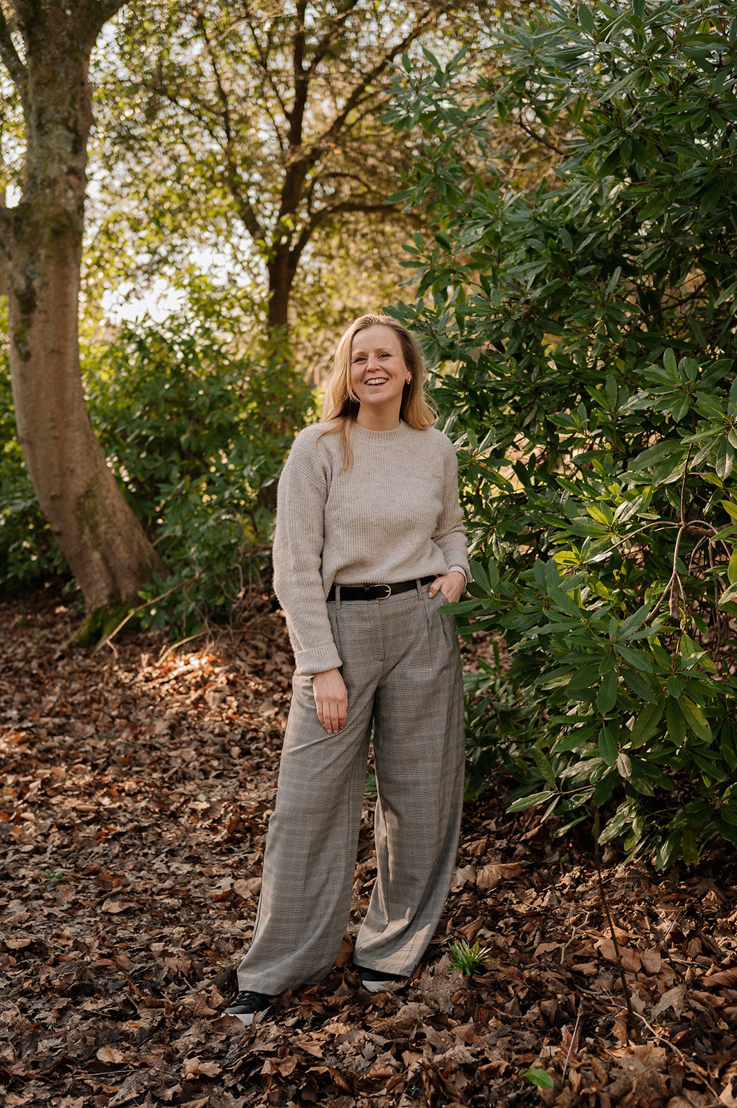 A person wearing a beige sweater and grey plaid trousers smiles while standing amidst an outdoor scene with tall green shrubs and scattered dry leaves, highlighted by warm sunlight filtering through the trees.