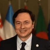 A smiling person with curly hair sits at a desk, looking at a laptop against a dark background.
