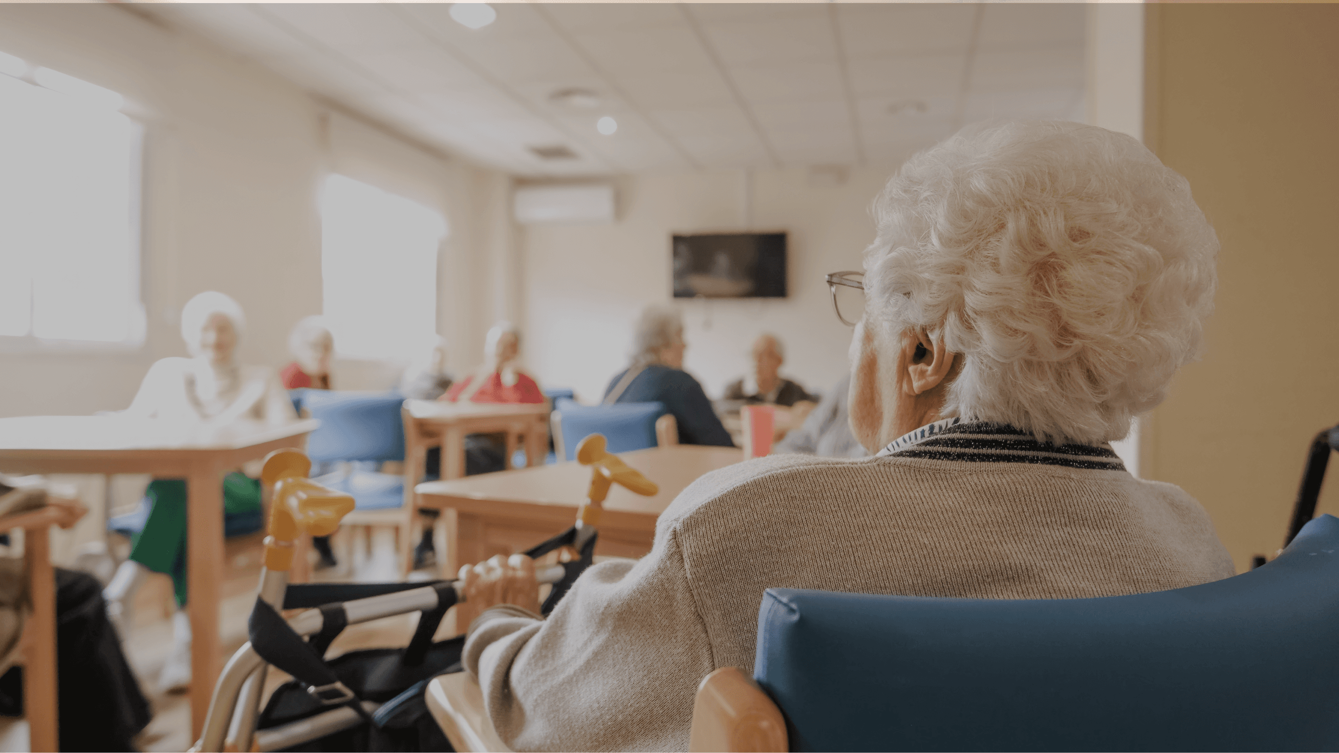 Elderly person seated in a care home lounge facing a group of other seniors socializing in the background.