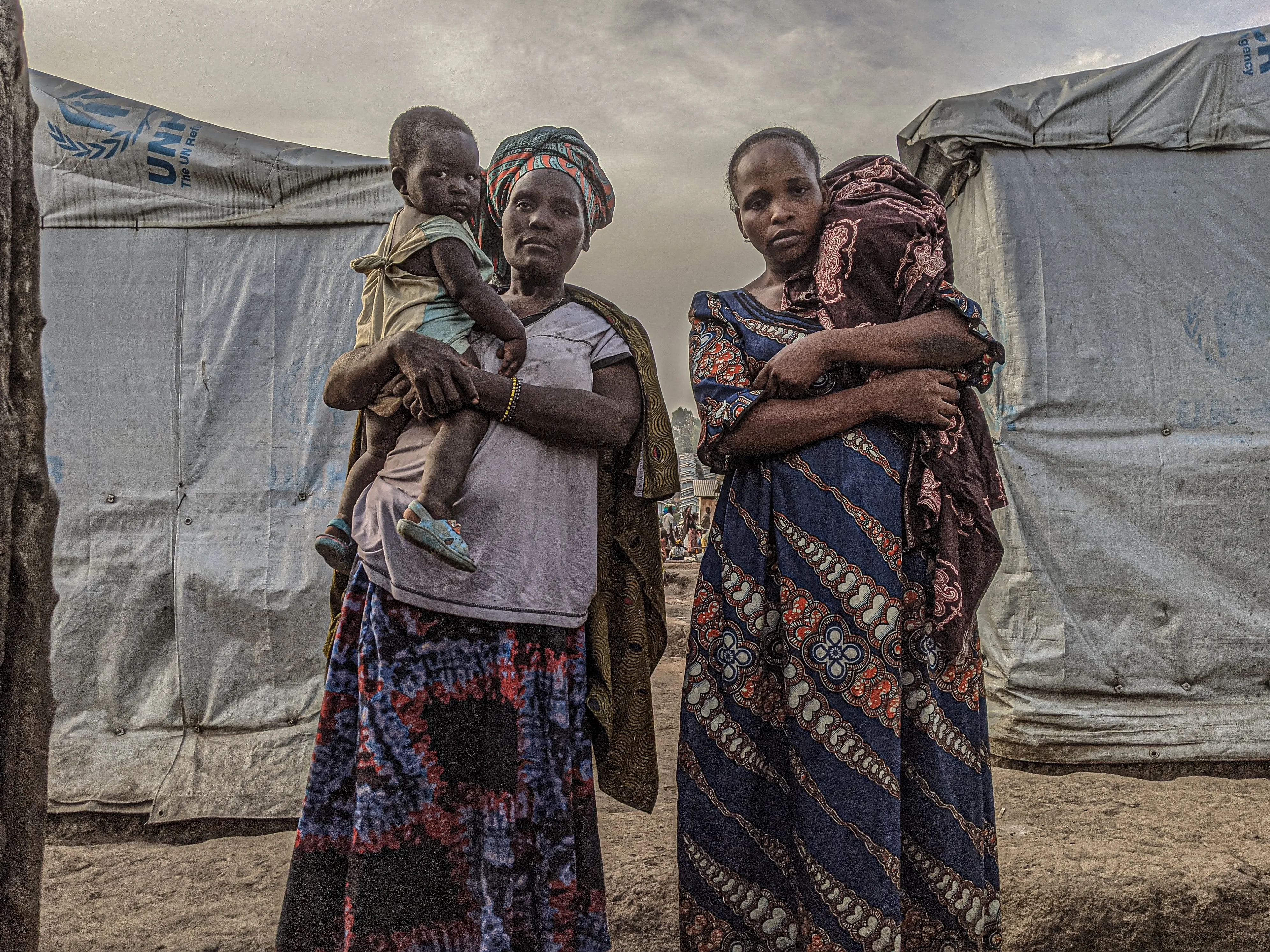 Two women who fled their former IDP camp because of insecurity, stand in front of their shelter in the Kigonze site. [Esdras Tsongo/Al Jazeera]