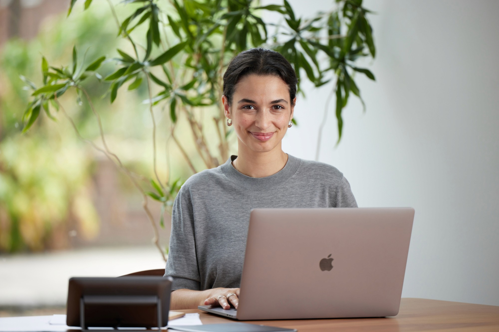 Woman smiling while working on a laptop at a desk in a bright workspace with indoor plants in the background.