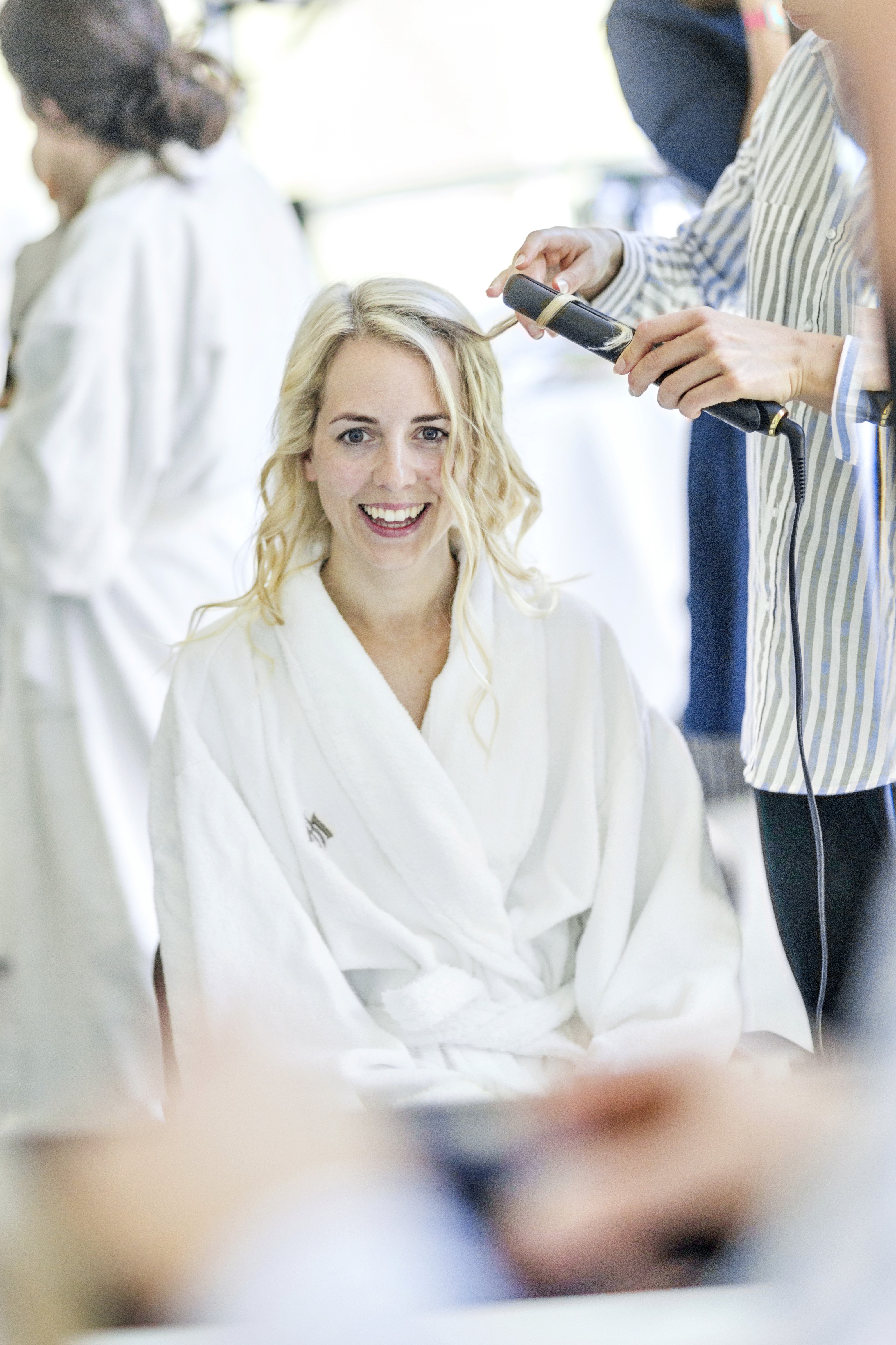 A woman in casual attire styles the hair of another woman in a white robe on a balcony, set against a backdrop of majestic mountains and a clear blue sky, suggesting a serene and intimate moment during a scenic getaway.