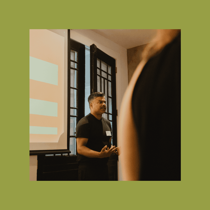 Man stands confidently during a presentation, facing an audience. He gestures expressively against a backdrop of a screen and windows, suggesting engagement.