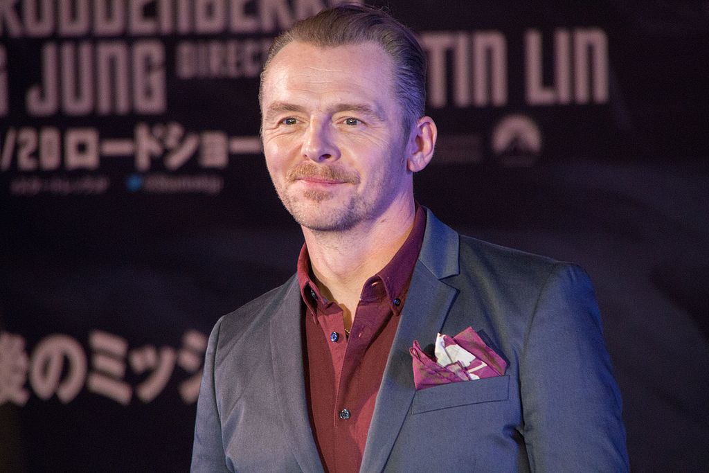 Simon Pegg in a grey suit with a red shirt and pocket square stands in front of a backdrop with text. He has short hair and a mustache, smiling slightly. The background features mixed-language text and logos.