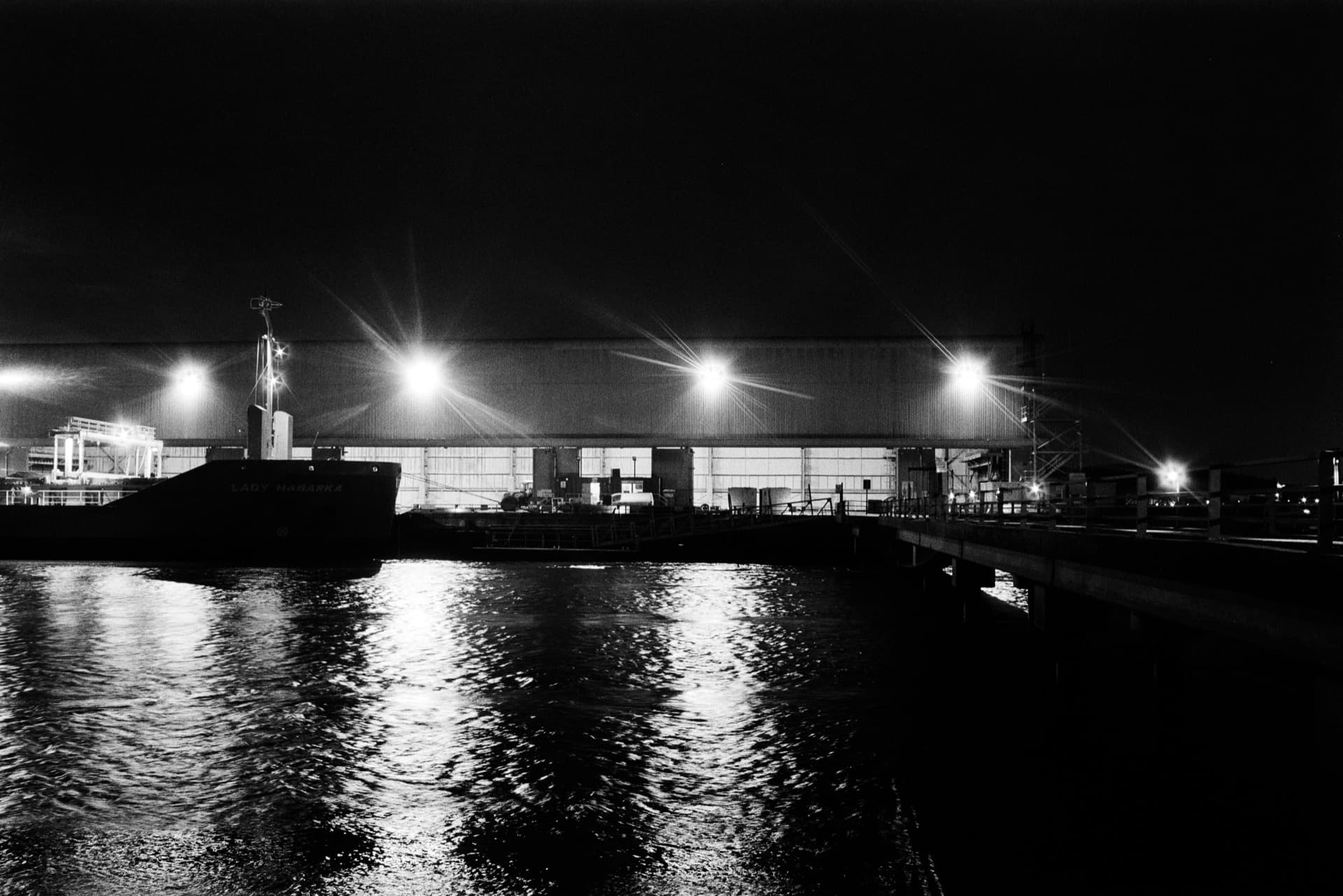 Moored vessel and illuminated industrial warehouse beside water at night with light reflections