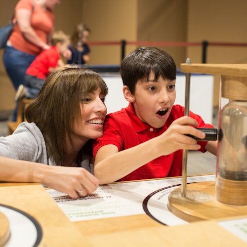 Una mujer sonriente y un niño emocionado interactúan con una exhibición práctica en un museo, con otras personas de fondo.