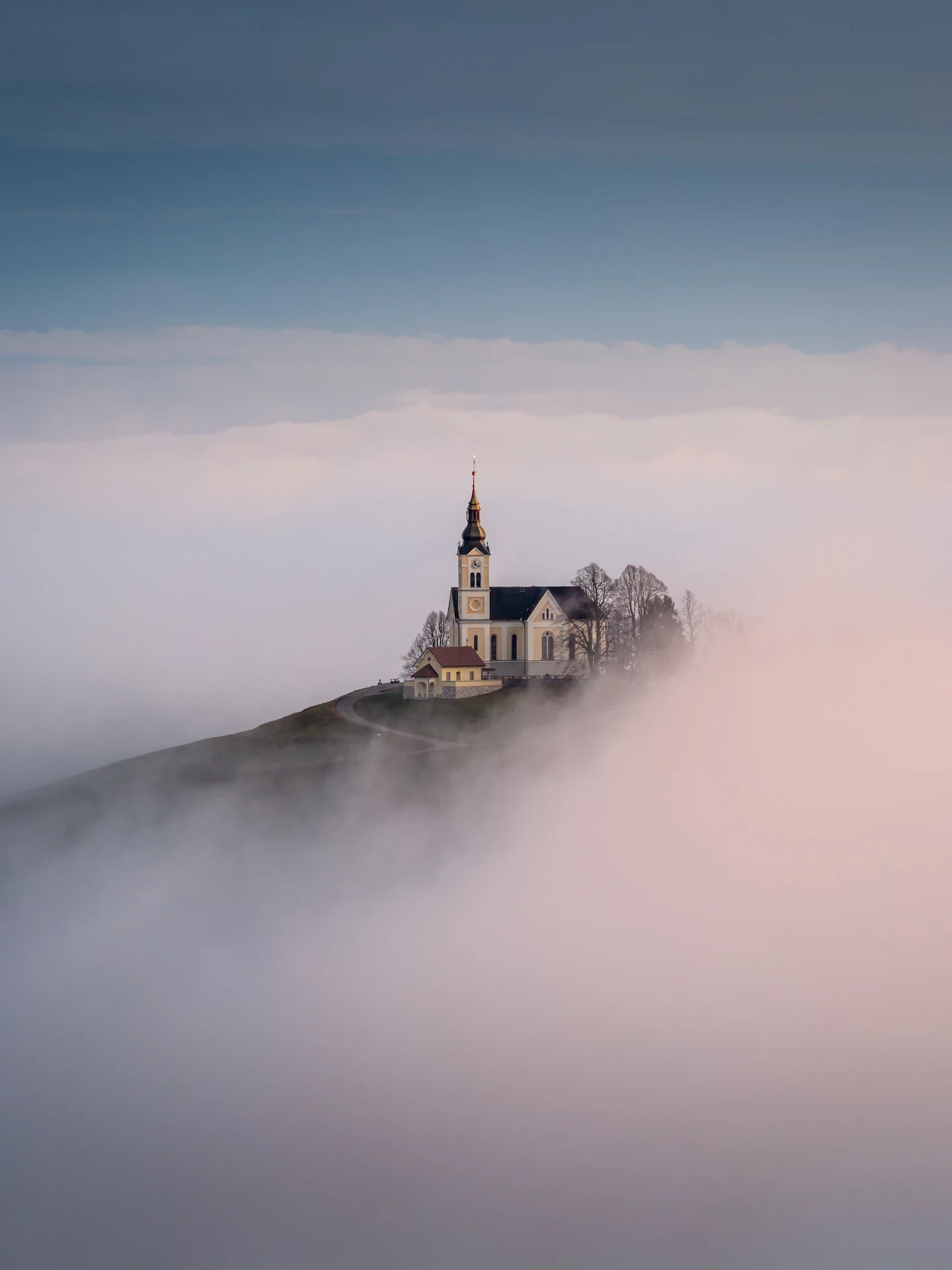 A tranquil scene shows a church in Slovenia emerging from dense fog against a soft gradient sky.