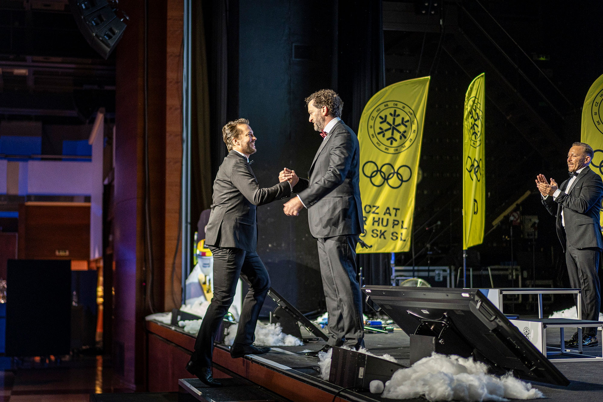 Two men in tuxedos shaking hands on stage during the gala evening at an international corporate conference, with Olympic-themed branding in the background.