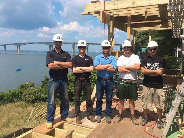 A group of five people in hard hats poses on a construction site with a bridge and water in the background.