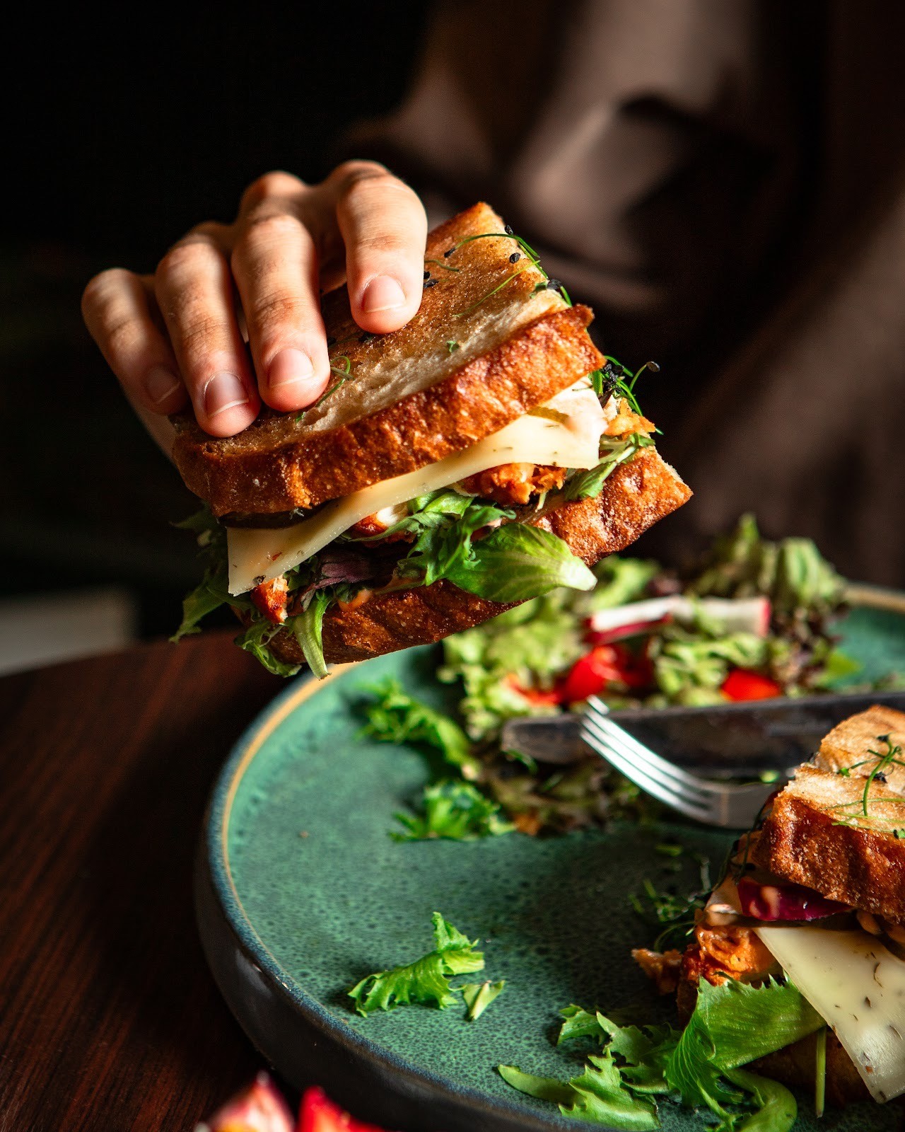 Nahaufnahme einer Hand, die ein köstlich aussehendes gegrilltes Sandwich mit Käse, Salat und einer herzhaften Füllung neben einem kleinen Beilagensalat hält.