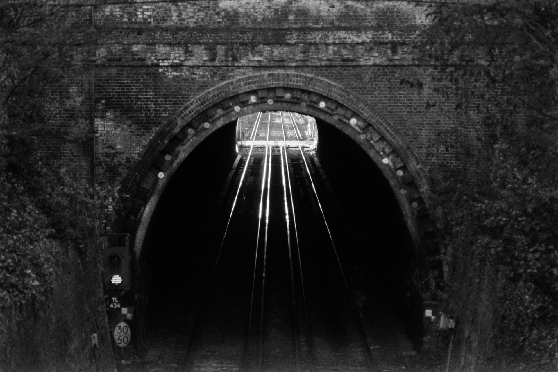 Railway tunnel entrance framed by weathered brick arch, converging rail lines leading through to bright exit