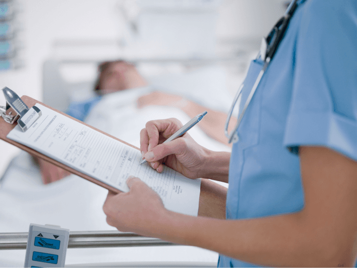 Nurse writing notes on clipboard beside patient in hospital bed.