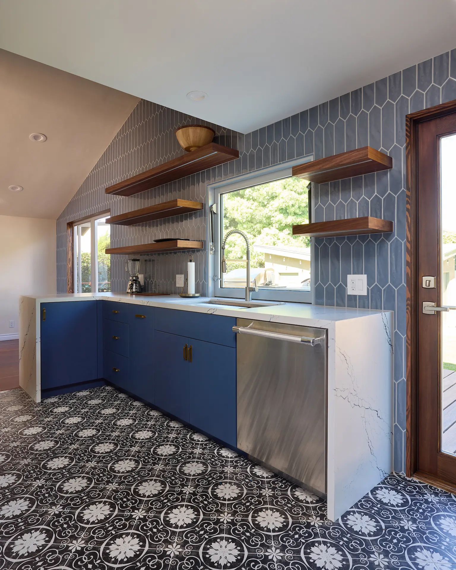 Portrait at an angle showing the entire sink wall with blue tile backsplash, cabinetry, and brown wooden floating shelves with LED under lighting, highlighting the functional elegance of the Huntington Beach Kitchen. Photo by Todd Huge.