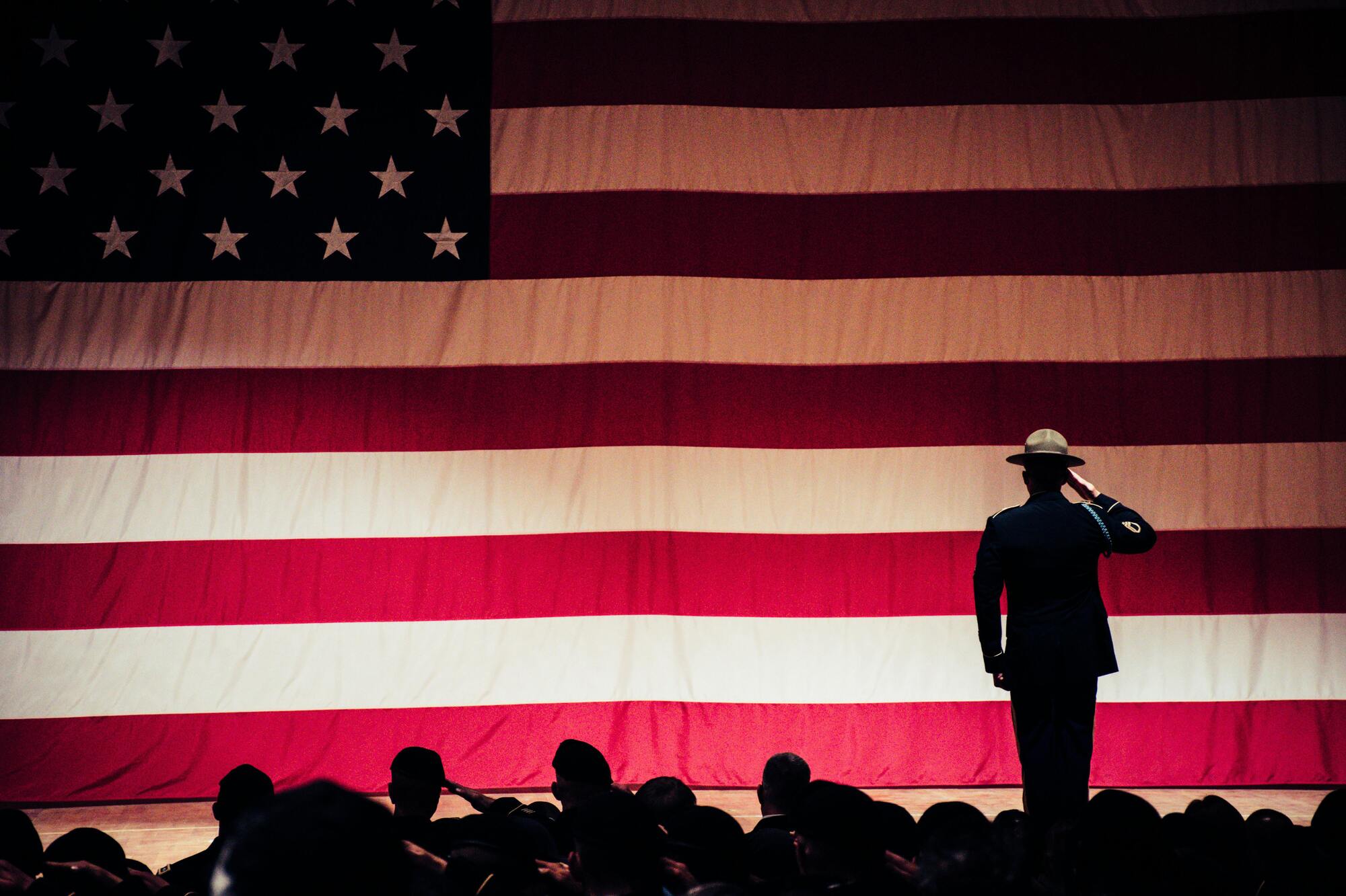 A solemn figure stands in salute before a large American flag draped in the background, symbolizing patriotism and respect.