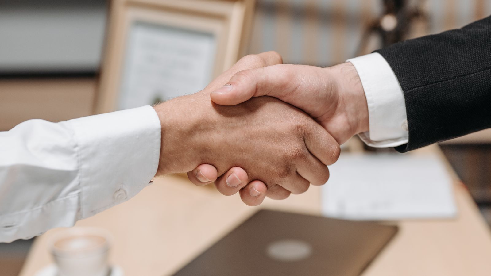 Two professionals shaking hands over a desk after agreeing terms, a laptop and document nearby
