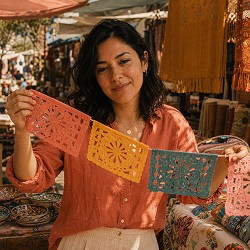 Woman in a vibrant market holding colorful papel picado banners in pink, yellow, and blue. She smiles gently, surrounded by crafts and textiles.