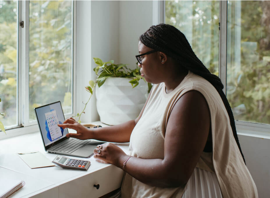 A woman in a beige dress and eye glasses working on her laptop in a windowed office content with her eb1 lawyers.