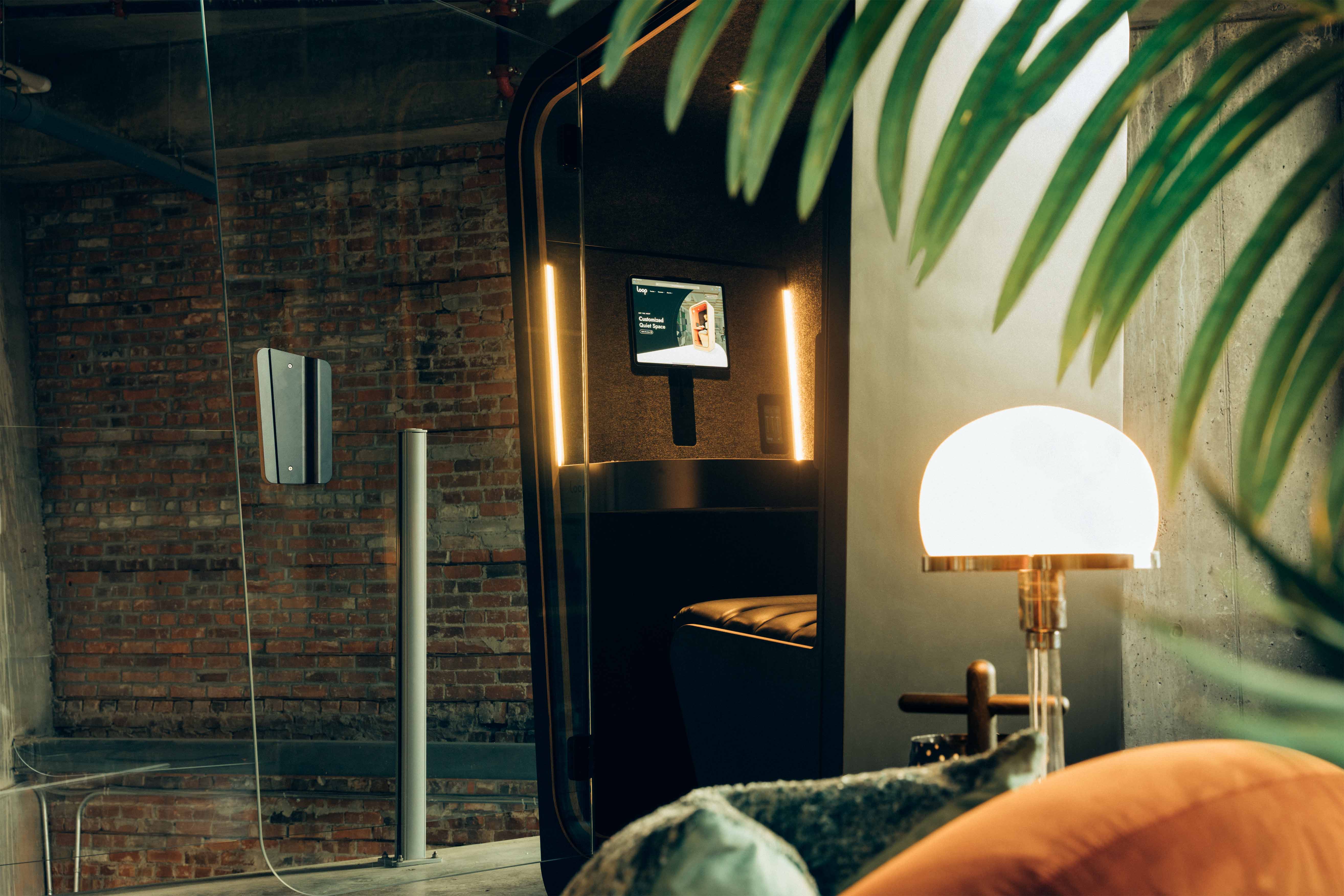 a privacy pod partially blocked by a plant in a modern office with exposed brick walls