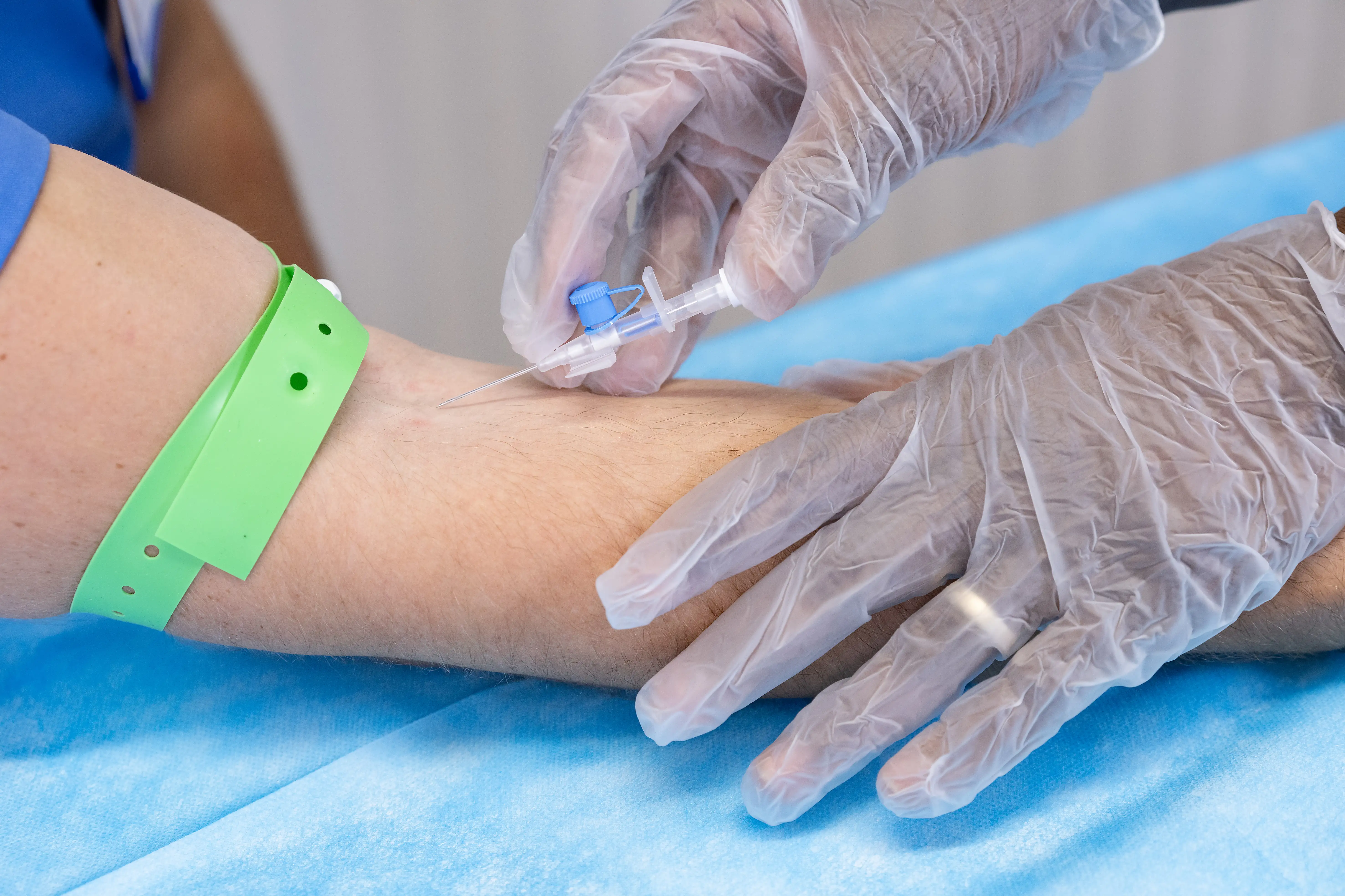 Healthcare professional wearing gloves inserting an IV cannula into a patient’s arm, with a green tourniquet applied and sterile blue drape underneath.