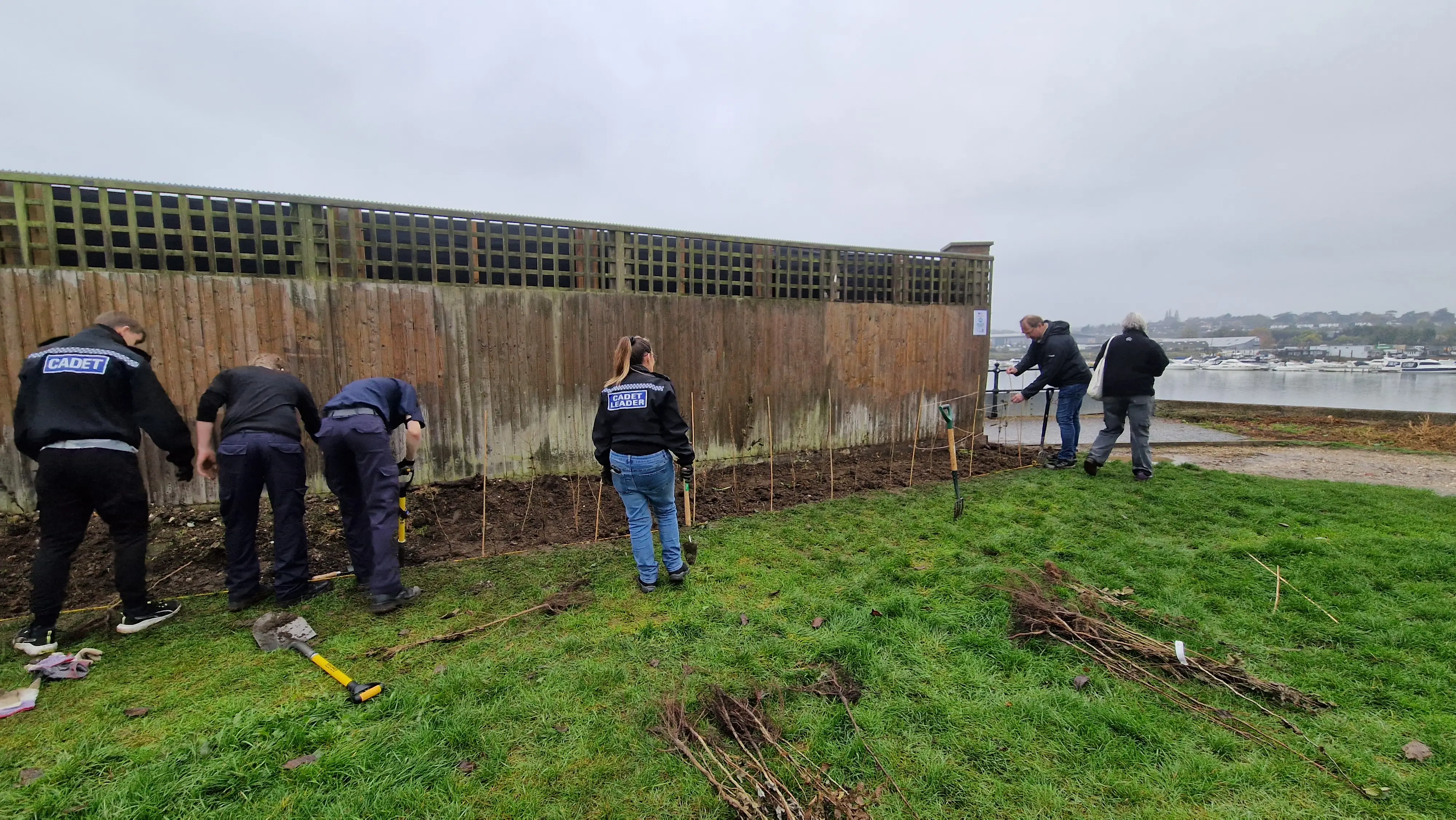 A group of people working near a wooden fence, with some standing on grass and others focused on a task.
