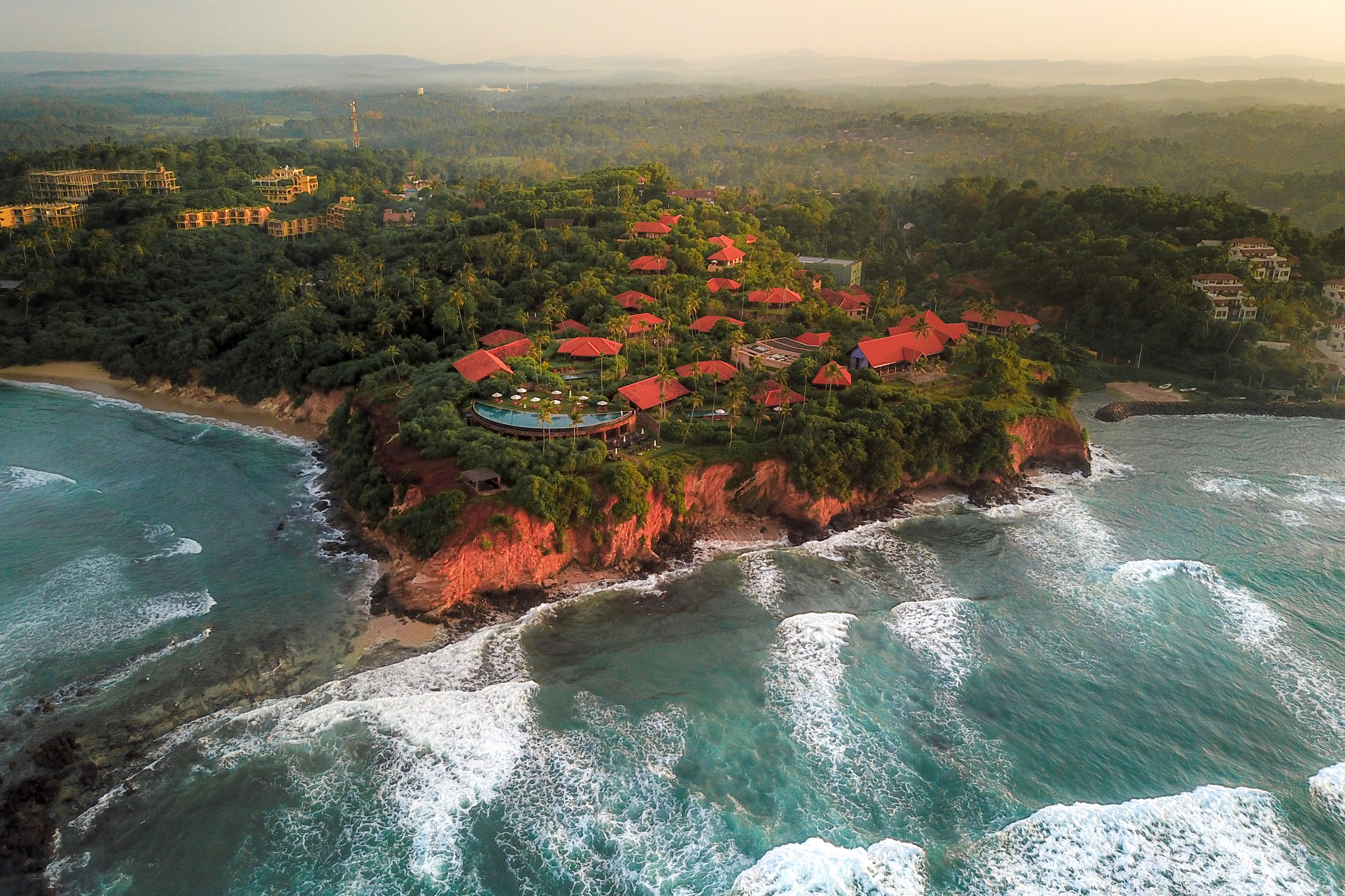 Aerial view of a coastal resort with red-roofed buildings nestled in green trees on a rocky cliff overlooking turquoise