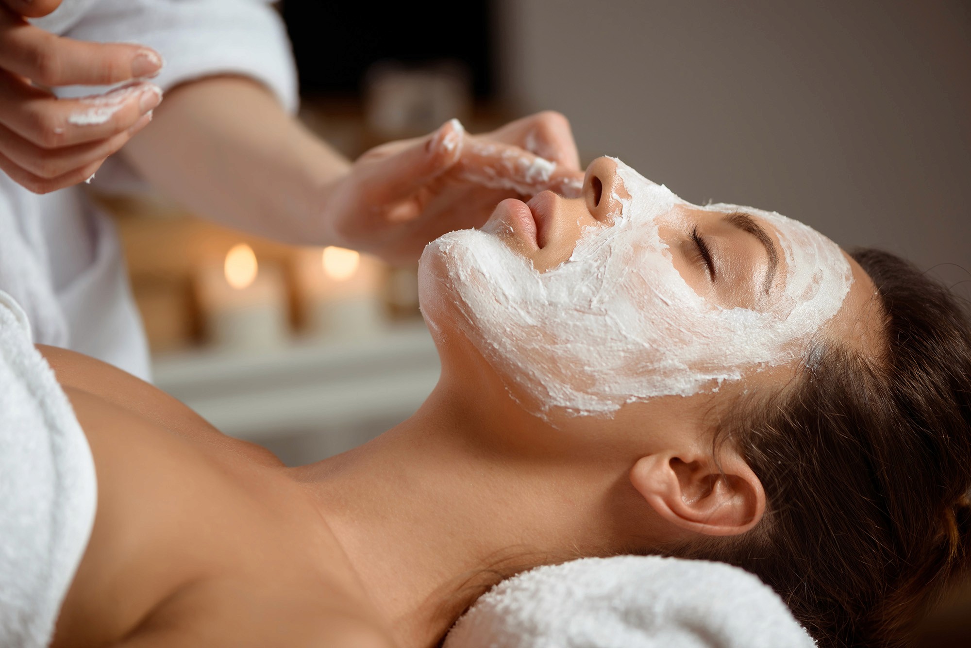 Woman lying down with a white facial mask being applied, surrounded by a calming spa environment