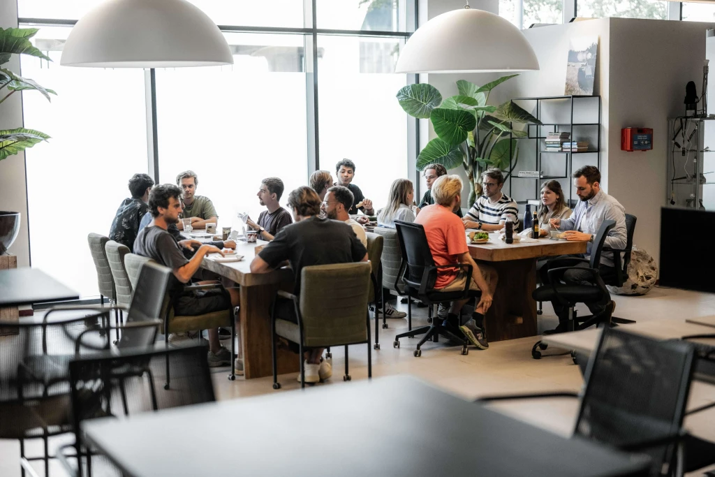 A group of people are gathered around a long wooden conference table in a modern office setting, with large windows and indoor plants enhancing the open, collaborative atmosphere.