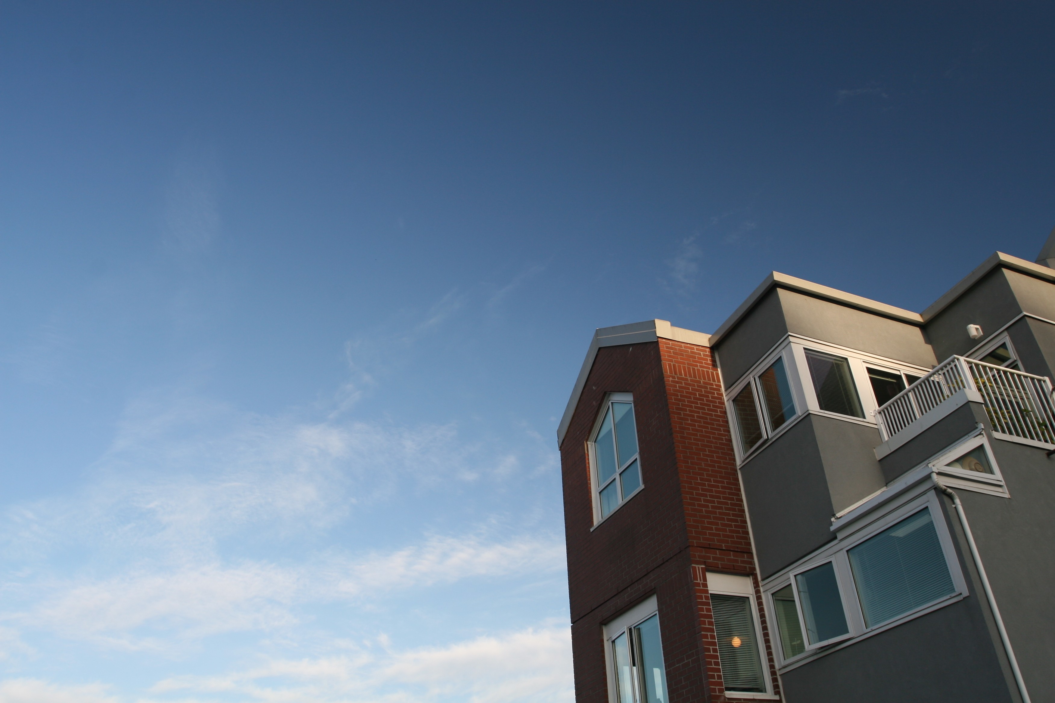 Exterior of a modern multifamily residential building at dusk, featuring a blue-grey panel facade, flat and shed rooflines with a dark metal standing-seam accent, and warmly lit windows against a deep blue gradient sky.