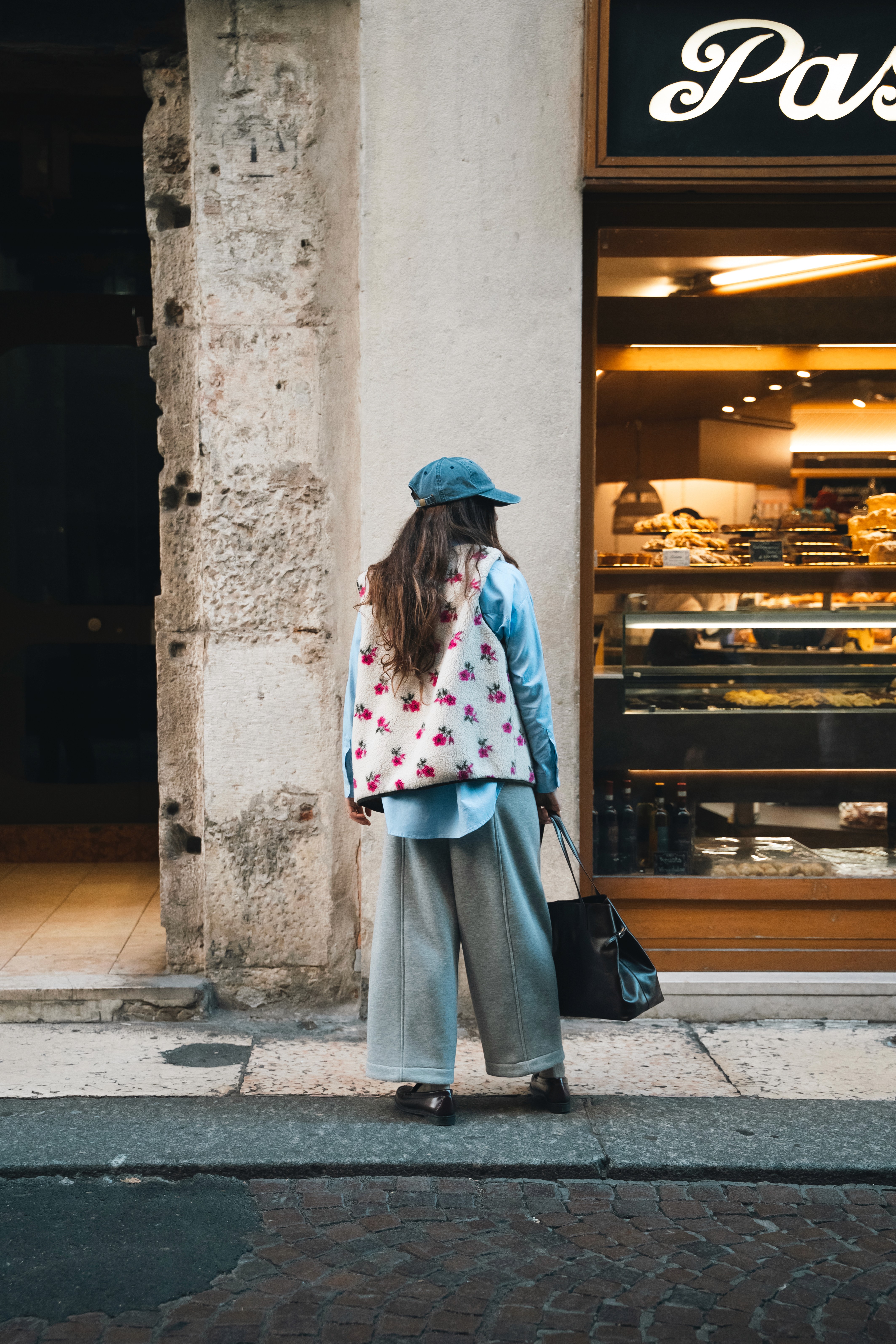 Une femme vue de dos dans une rue Italienne avec un gilet à motifs, un pantalon large et une chemise bleue dans le lookbook de Paper Wardrobe