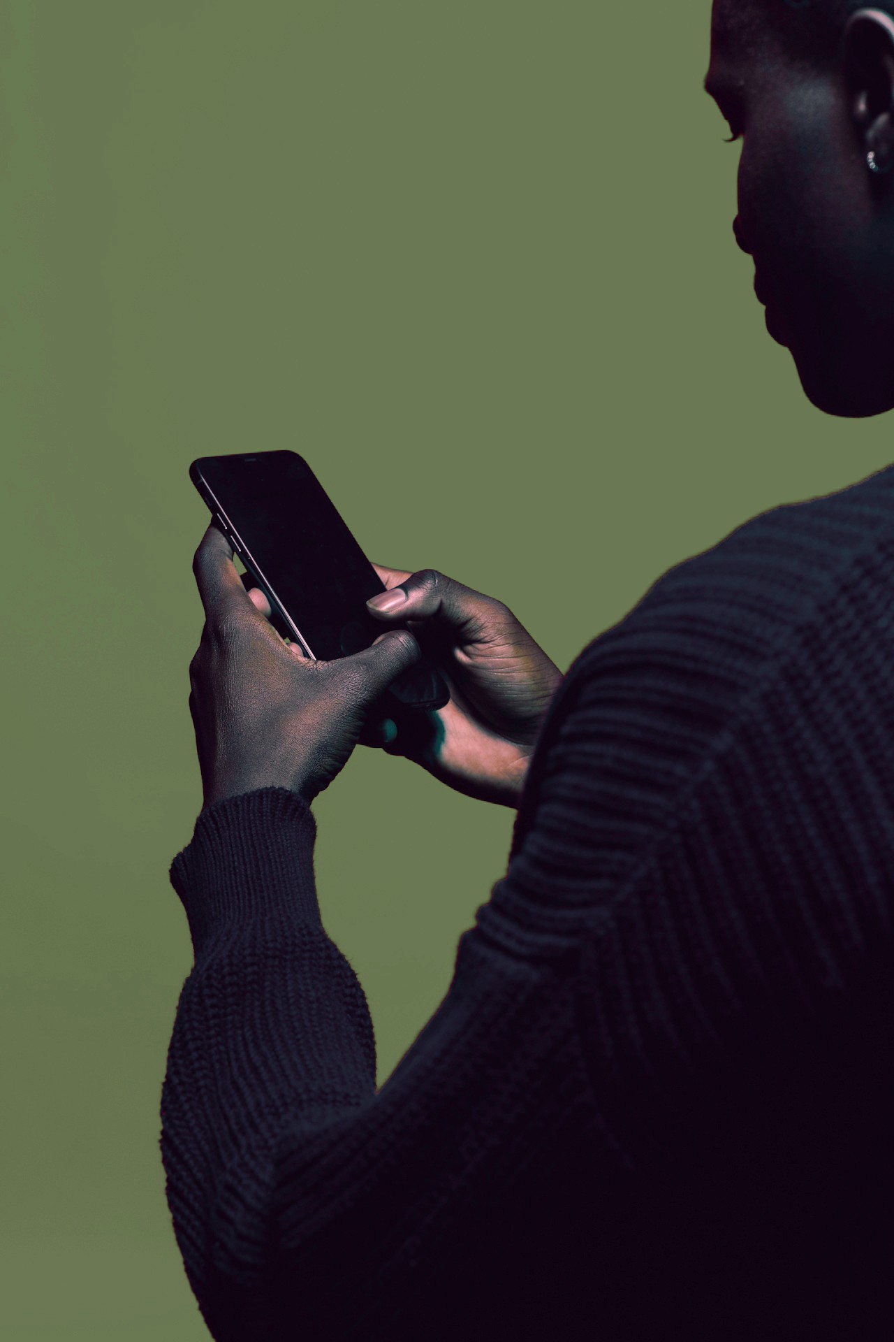 Eine Person hält ein Smartphone in den Händen, vor einem grünen Hintergrund. Die Person ist von hinten zu sehen und trägt dunkle Kleidung.