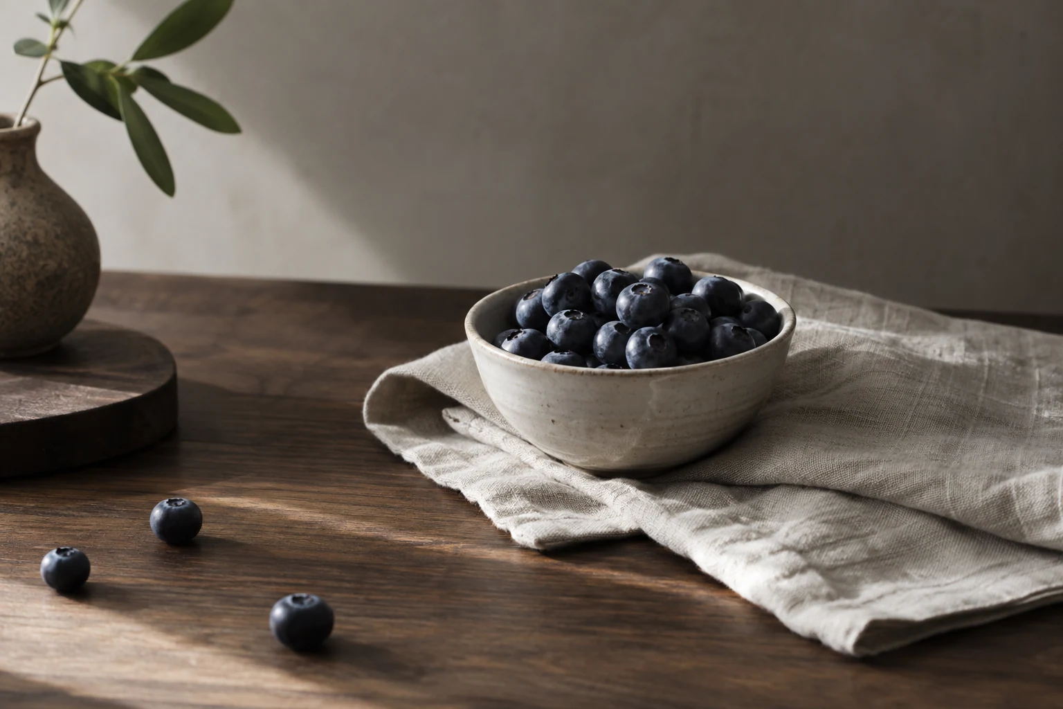 Small porcelain bowl of blueberries on folded pale linen on dark walnut, soft diagonal daylight