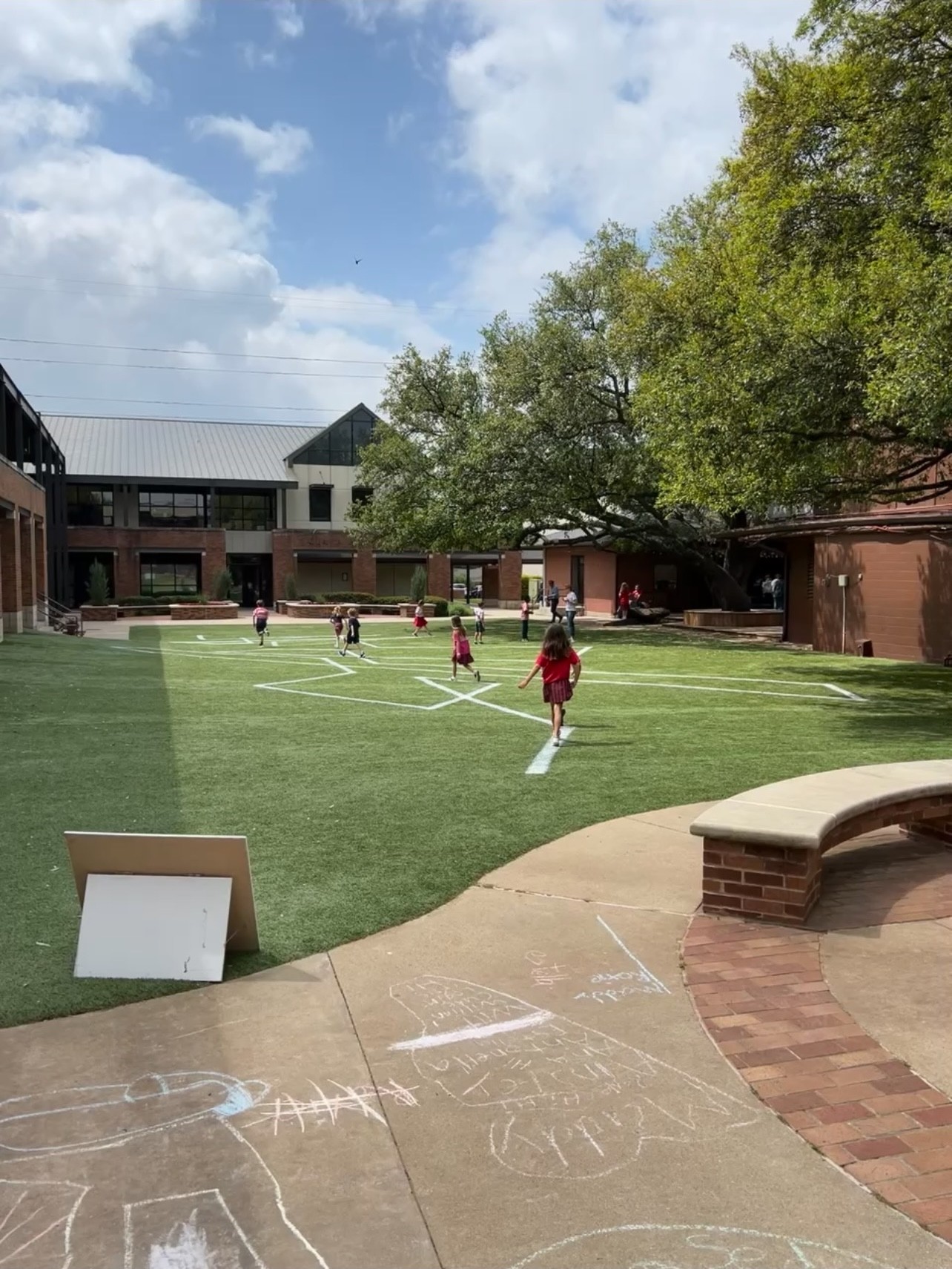 A sunny courtyard with grass, trees, and people enjoying outdoor activities. A seating area is visible in the foreground.