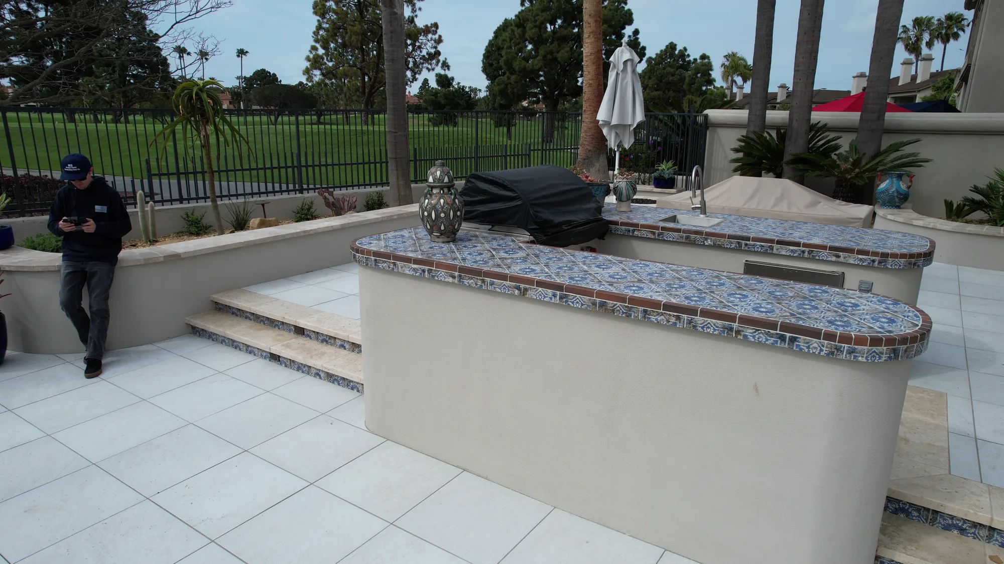 Outdoor kitchen with a patterned tile counter, covered barbecue and surrounding pale paving.