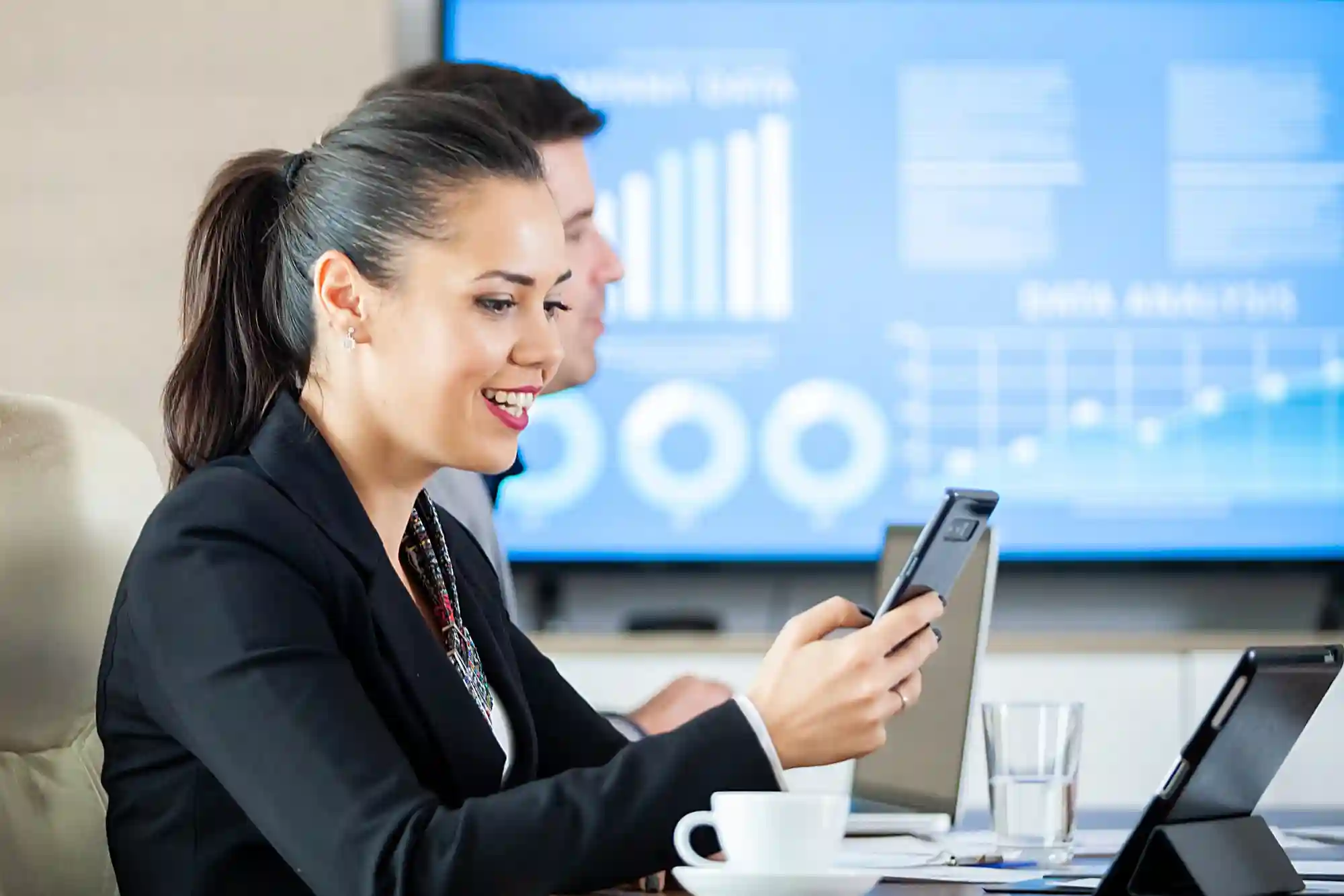 A professional woman smiling while using her smartphone in an office setting with data analytics displayed behind her.