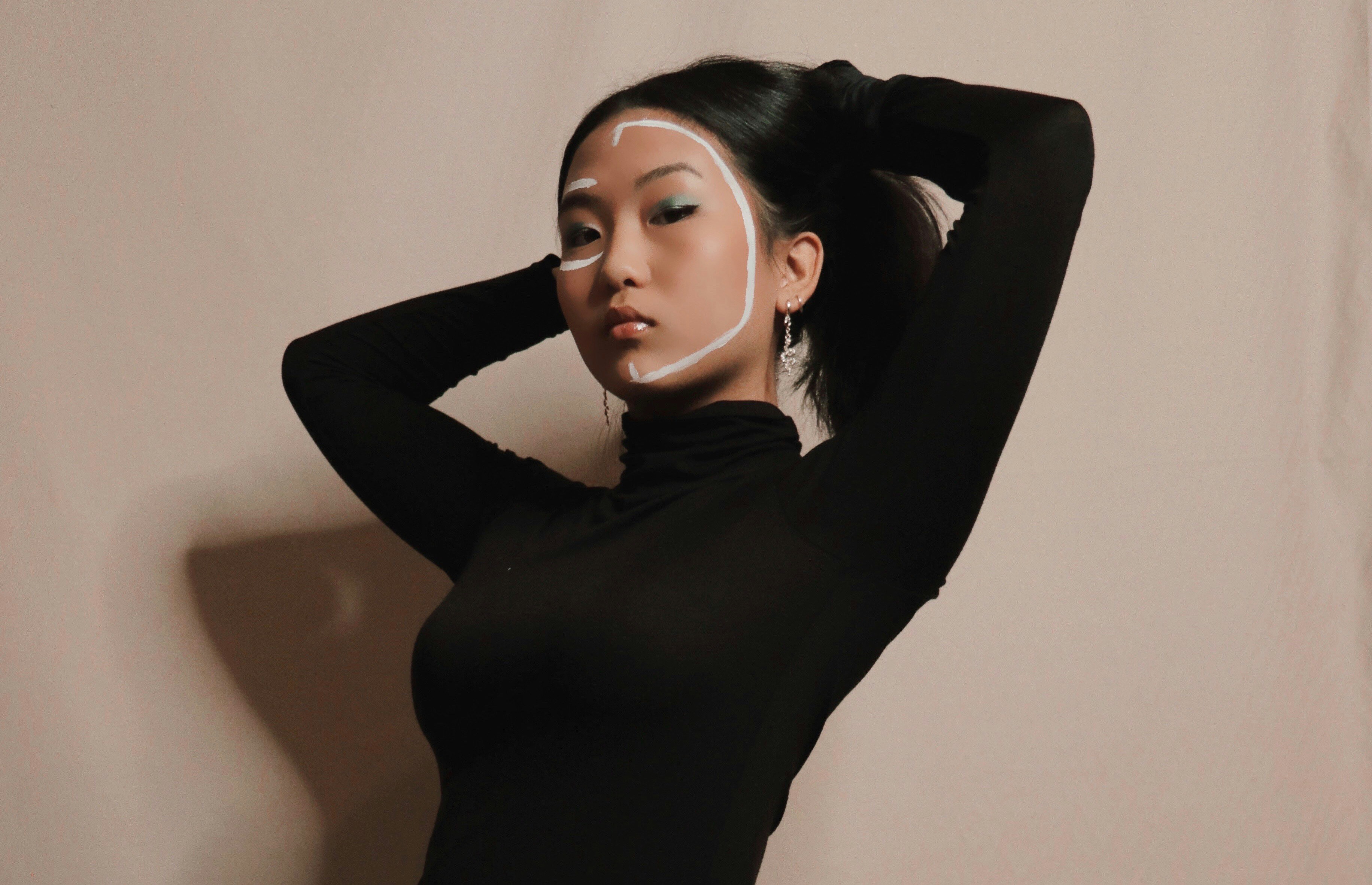 A woman in a black long-sleeve top poses with her arms raised, against a neutral background.