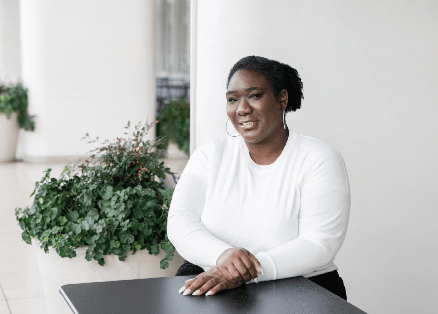 A medium shot captures therapist Sabrina Simpson with her hair pulled back, wearing a white long-sleeved shirt and l Sabrina Simpson is seated at a dark table, with her hands resting on its surface. Sabrina Simpson is looking to her right and smiling. Behind her, to the left, is a large planter filled with lush green foliage, and further back, a white pillar and a blurred background of more greenery and architectural elements. The lighting is soft and natural, creating a bright and airy atmosphere.