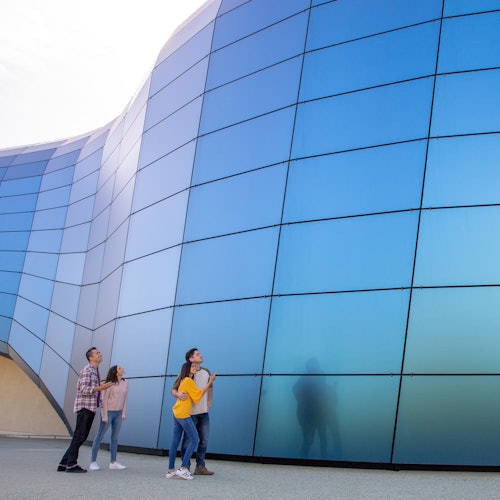 四个人站着仰望一座带有蓝色反光板的大型弧形建筑。