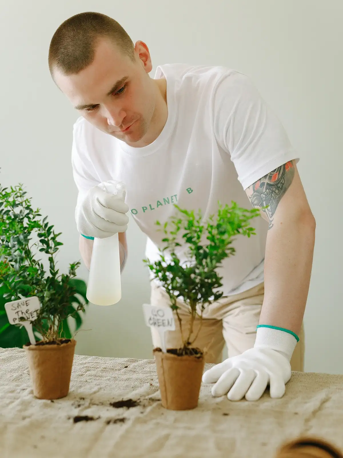 A person in a white shirt and gloves sprays water on potted plants labeled "Save the Planet" and "Go Green".