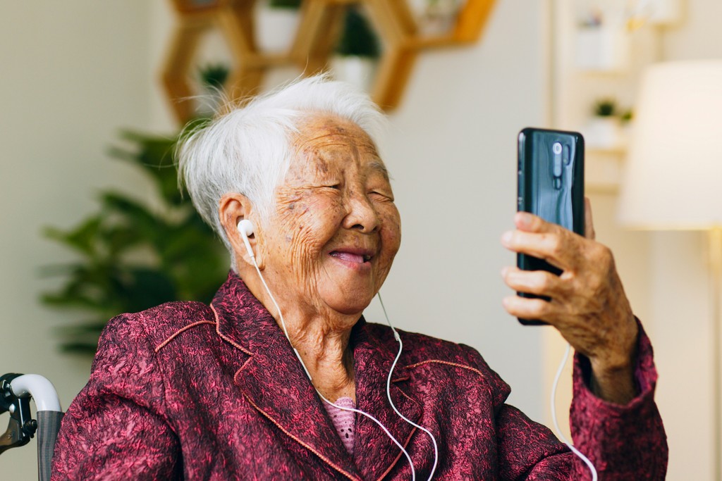 An elderly woman with earphones smiles warmly while holding a smartphone, enjoying a video call, highlighting the joy of staying connected with loved ones through modern technology.