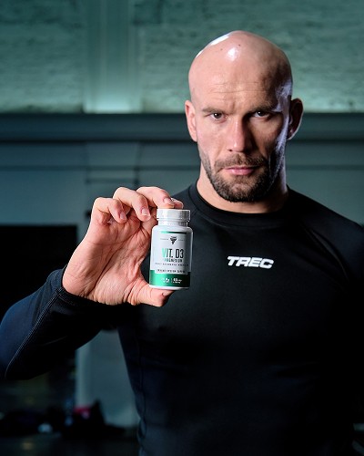 Fotografia produktowa / reklamowa przedstawiająca mężczyznę w koszulce TREC trzymającego opakowanie suplementu VIT. D3 + magnesium firmy TREC