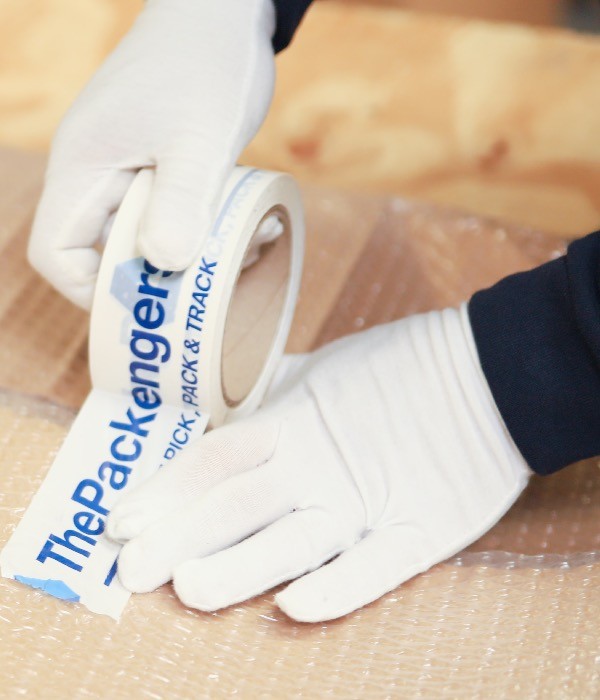 lose-up of a professional wearing white gloves and using branded ThePackengers tape to secure a fragile shipment, showcasing white-glove packaging expertise.