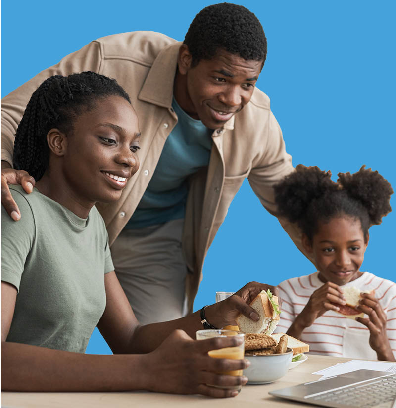 Family gathered around a table with a laptop and food, with adults and a child looking toward the screen, against a blue background.