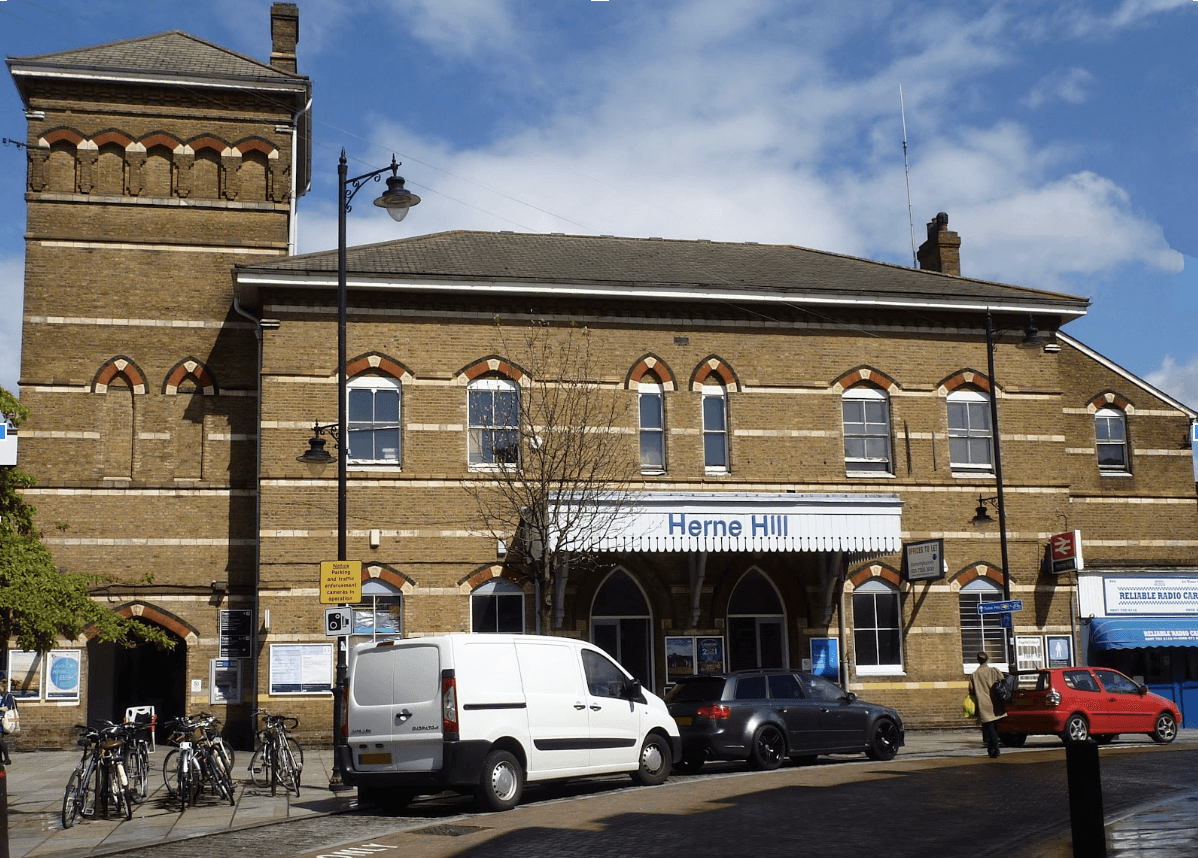 A smiling man with curly hair is sitting at a desk, looking at the camera, with a laptop and a lamp in the background.
