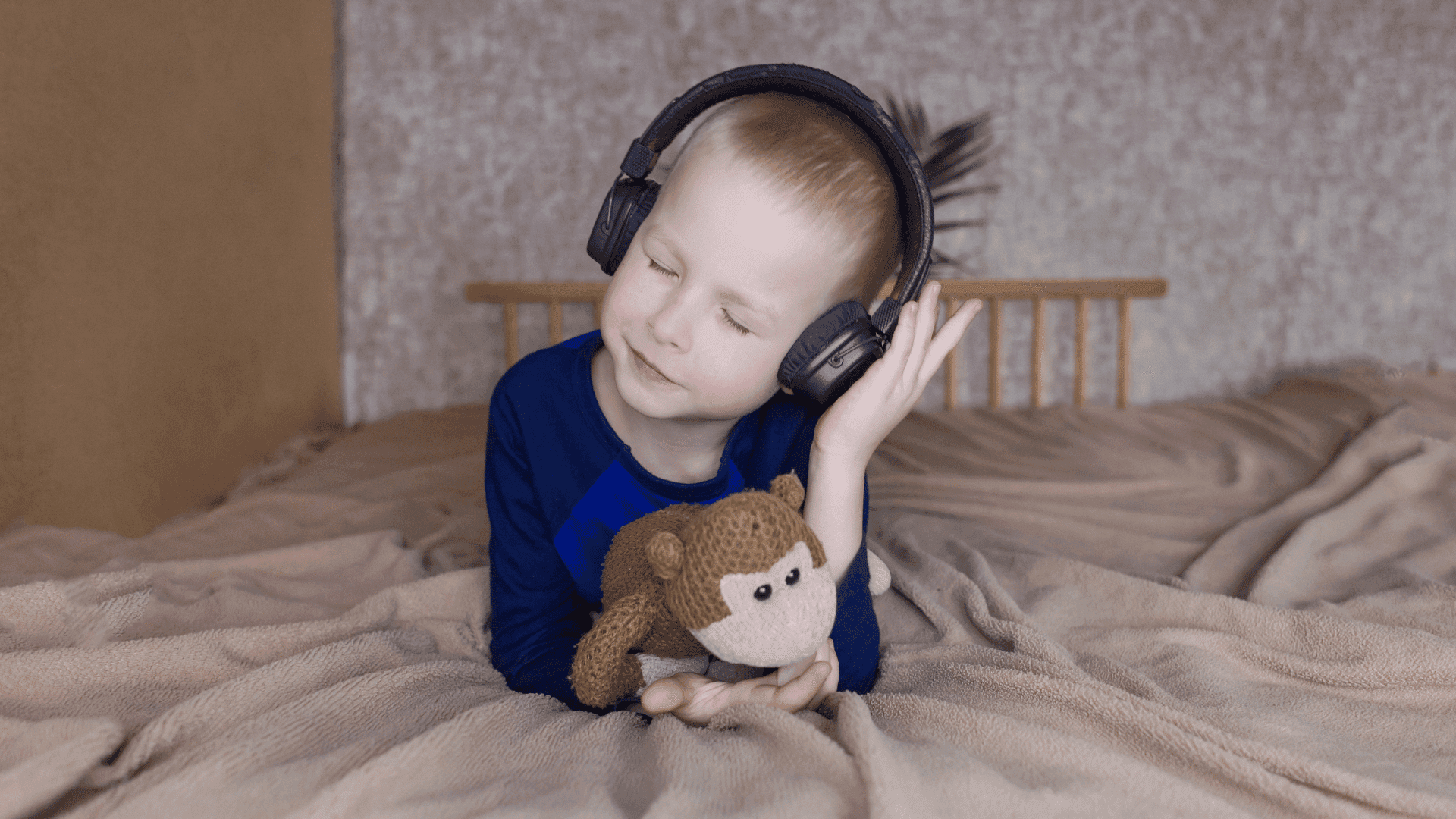 A young boy wearing a blue shirt lying on a bed, wearing headphones and holding a toy monkey while listening to HushAway®’s Sound Sanctuary.
