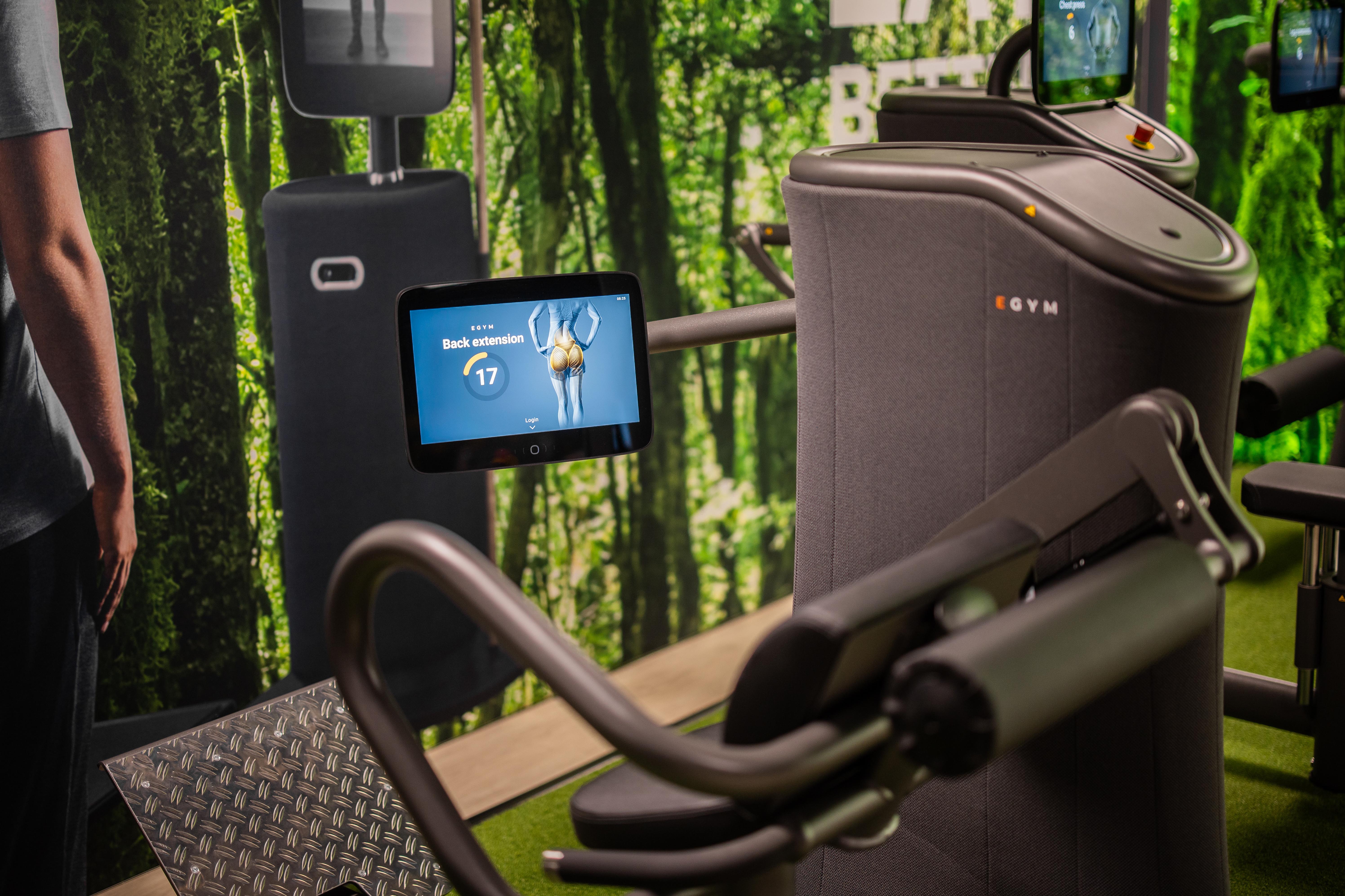 A close-up of exercise equipment in a fitness setting surrounded by greenery, with digital screens visible.