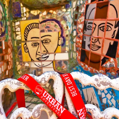 Mosaic art wall featuring colorful portraits and a foreground of a white, wavy sculpture holding a red band labeled "Urban Adventures."