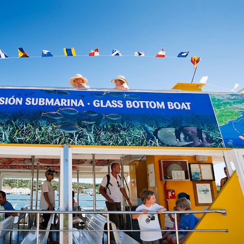 Glass-bottom boat with passengers, a colorful sign, and flags above. People onboard enjoy the ride on a sunny day.