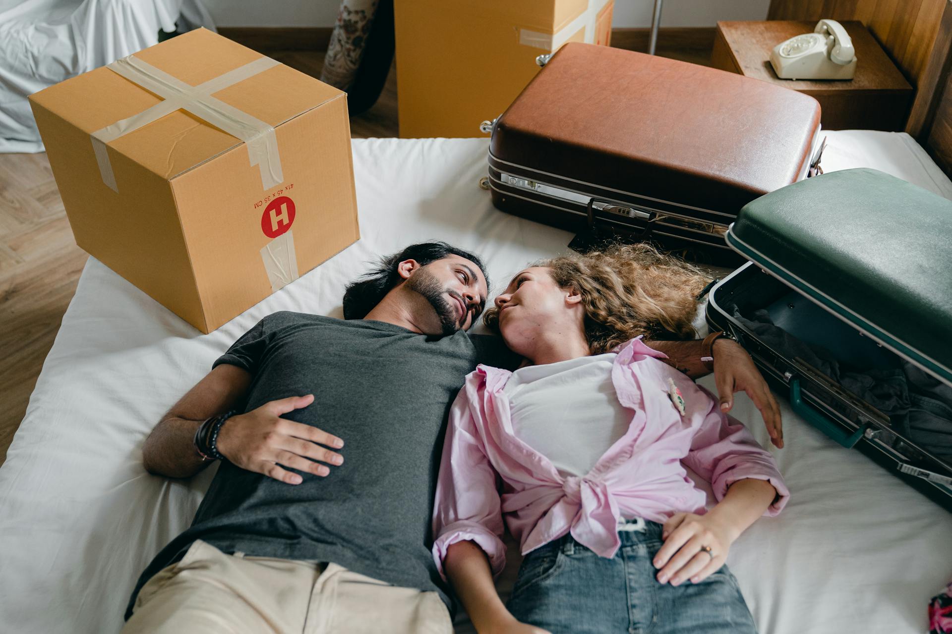  A couple is lying on a bed surrounded by moving boxes and suitcases, looking lovingly into each other's eyes. The man has long dark hair and a beard, and the woman has curly blonde hair, wearing a pink shirt tied at the waist.